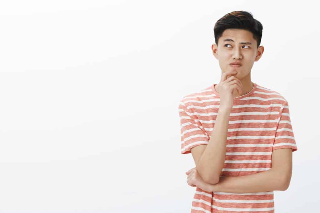 portrait troubled young asian guy trying think up plan idea standing thoughtful pose with hand chin looking questioned hesitant upper left corner